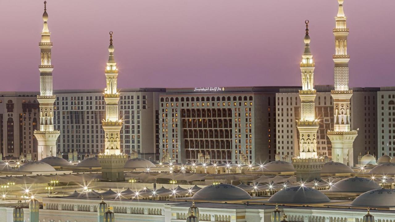 Hotel Intercontinental Dar Al Iman Madinah By Ihg Medyna Zewnętrze zdjęcie The photo depicts an architectural scene, likely from a mosque complex, featuring prominent minarets that are illuminated. The setting appears to be during twilight, with a soft gradient in the sky. In the background, a large building is visible, pos