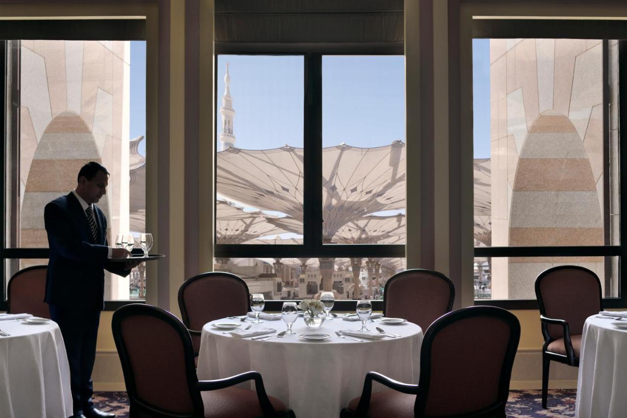 Hotel Intercontinental Dar Al Iman Madinah By Ihg Medyna Zewnętrze zdjęcie The photo shows a dining area with elegant decor. A waiter is standing to the side, holding a tray. The tables are neatly set with white tablecloths and fine dining arrangements. Large windows provide a view of an outdoor area featuring architectural