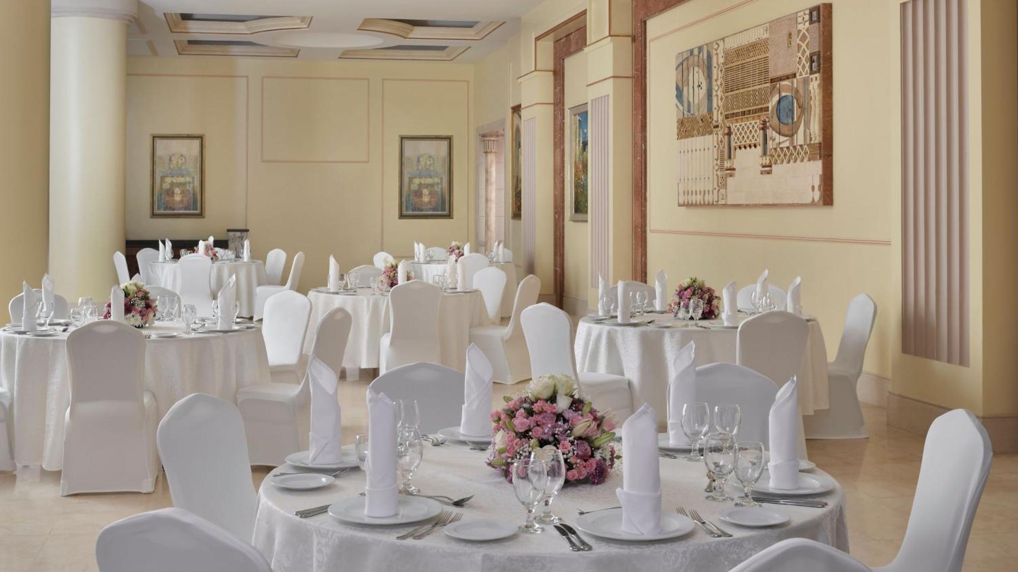 Hotel Intercontinental Dar Al Iman Madinah By Ihg Medyna Zewnętrze zdjęcie The photo shows an elegantly arranged dining area, likely set up for a formal event or reception. There are several round tables covered with white tablecloths, each adorned with floral centerpieces featuring pink flowers. The tables are set with pla