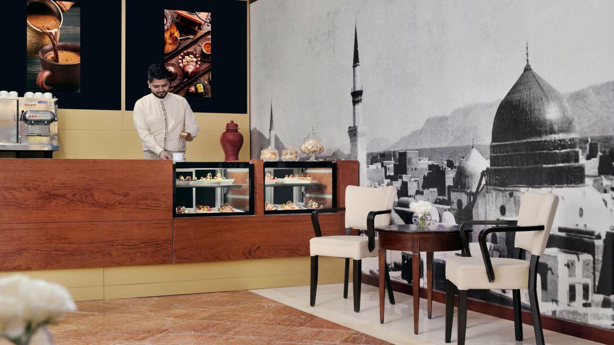 Hotel Intercontinental Dar Al Iman Madinah By Ihg Medyna Zewnętrze zdjęcie The photo shows an elegant cafe or dessert shop interior. A man in traditional attire is standing behind a wooden counter, where various pastries and sweets are displayed. The walls feature a large black-and-white mural of a cityscape, possibly depic