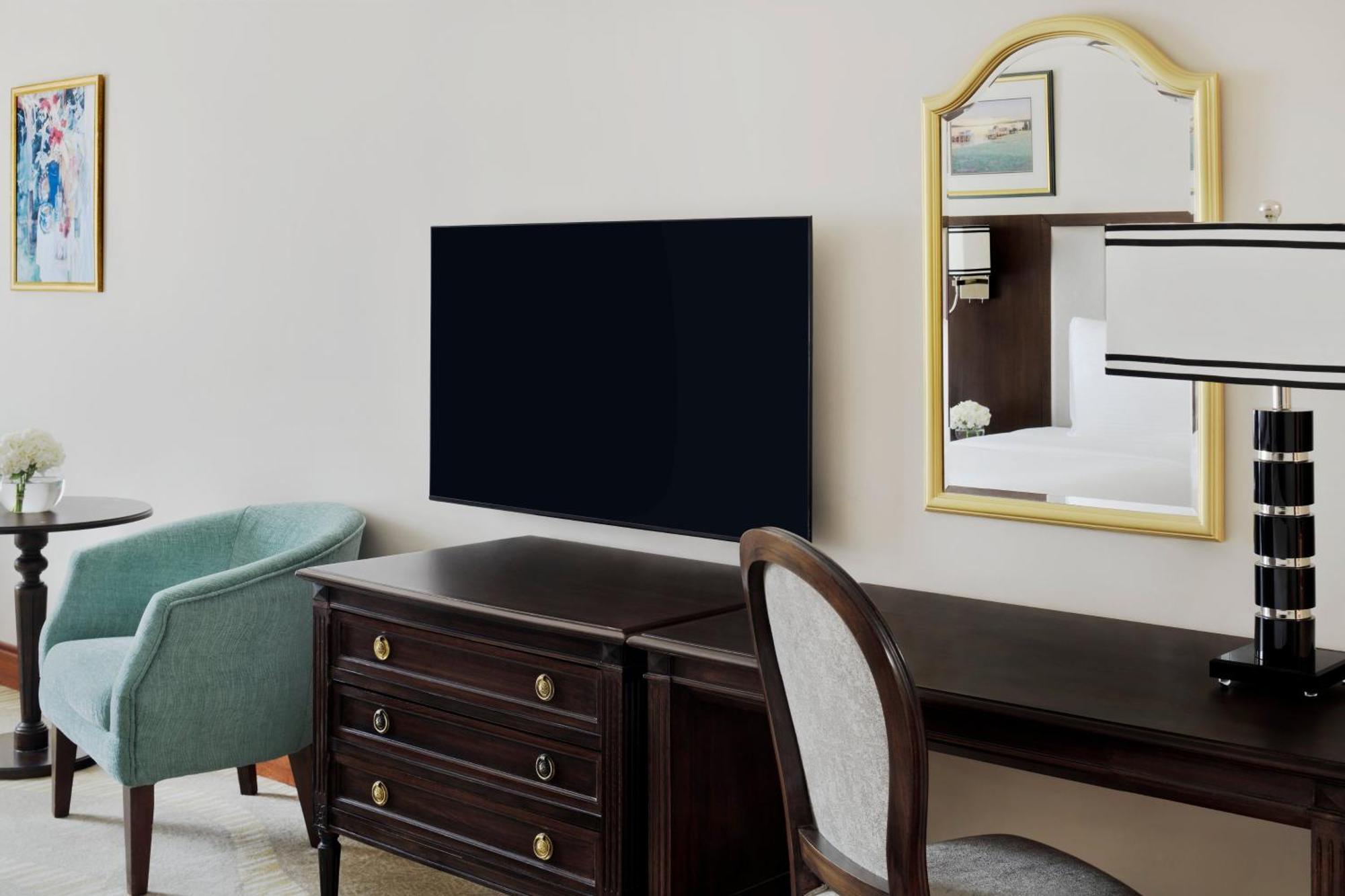 Hotel Intercontinental Dar Al Iman Madinah By Ihg Medyna Zewnętrze zdjęcie The photo shows a hotel room interior featuring a mounted flat-screen television on the wall. Below the TV, there is a dark wooden dresser with drawers. To the right, there's a wooden desk and a chair with a light-colored seat. A small table with a l
