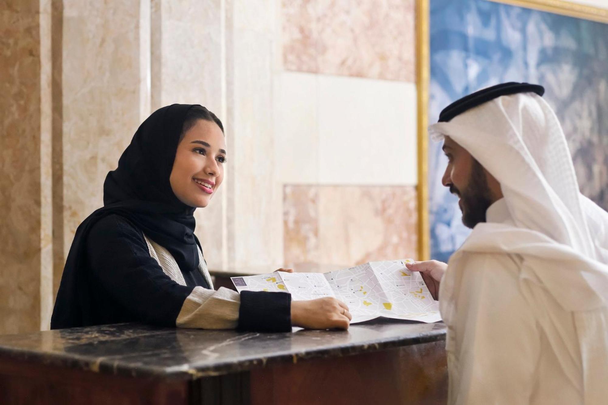 Hotel Intercontinental Dar Al Iman Madinah By Ihg Medyna Zewnętrze zdjęcie The photo depicts a woman in a black hijab and a light-colored outfit smiling as she interacts with a man wearing traditional Emirati attire, including a white kandura and ghutrah (headscarf). They are at a reception desk in what appears to be a hote