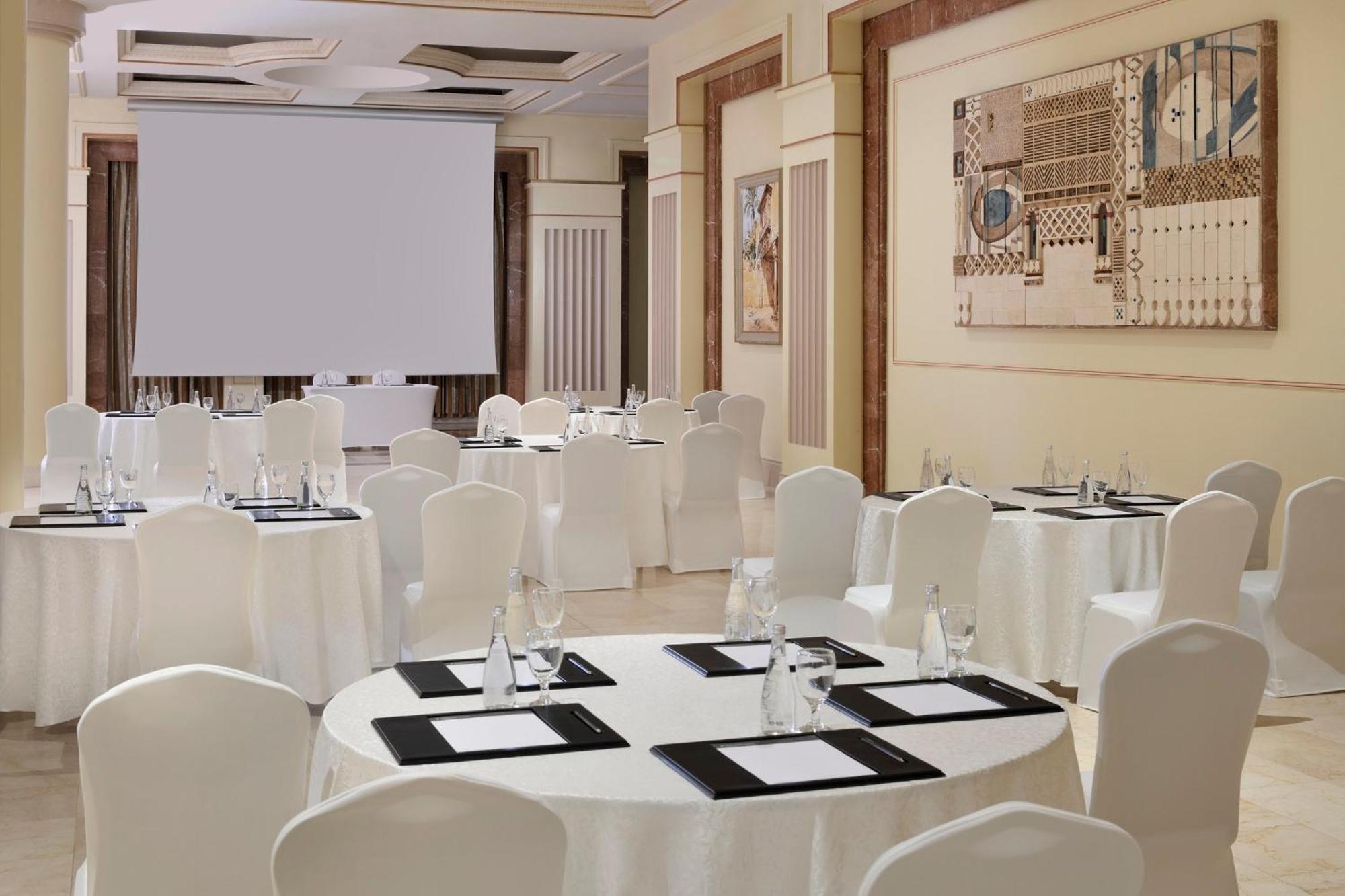 Hotel Intercontinental Dar Al Iman Madinah By Ihg Medyna Zewnętrze zdjęcie The photo shows a well-decorated event space or meeting room. It features several round tables covered with white tablecloths and surrounded by white chairs. Each table has black placemats and glass water bottles, suggesting a formal setup for a gath