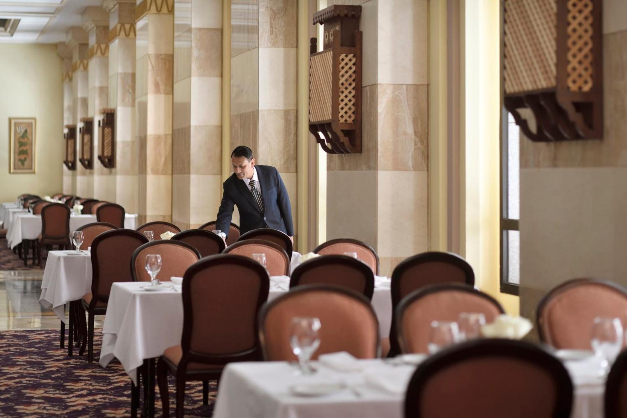 Hotel Intercontinental Dar Al Iman Madinah By Ihg Medyna Zewnętrze zdjęcie The photo depicts a dining area in an elegant restaurant. There are several tables set with white tablecloths, each accompanied by chairs. A waiter, dressed in a formal suit, is seen arranging or tending to one of the tables. The restaurant features 