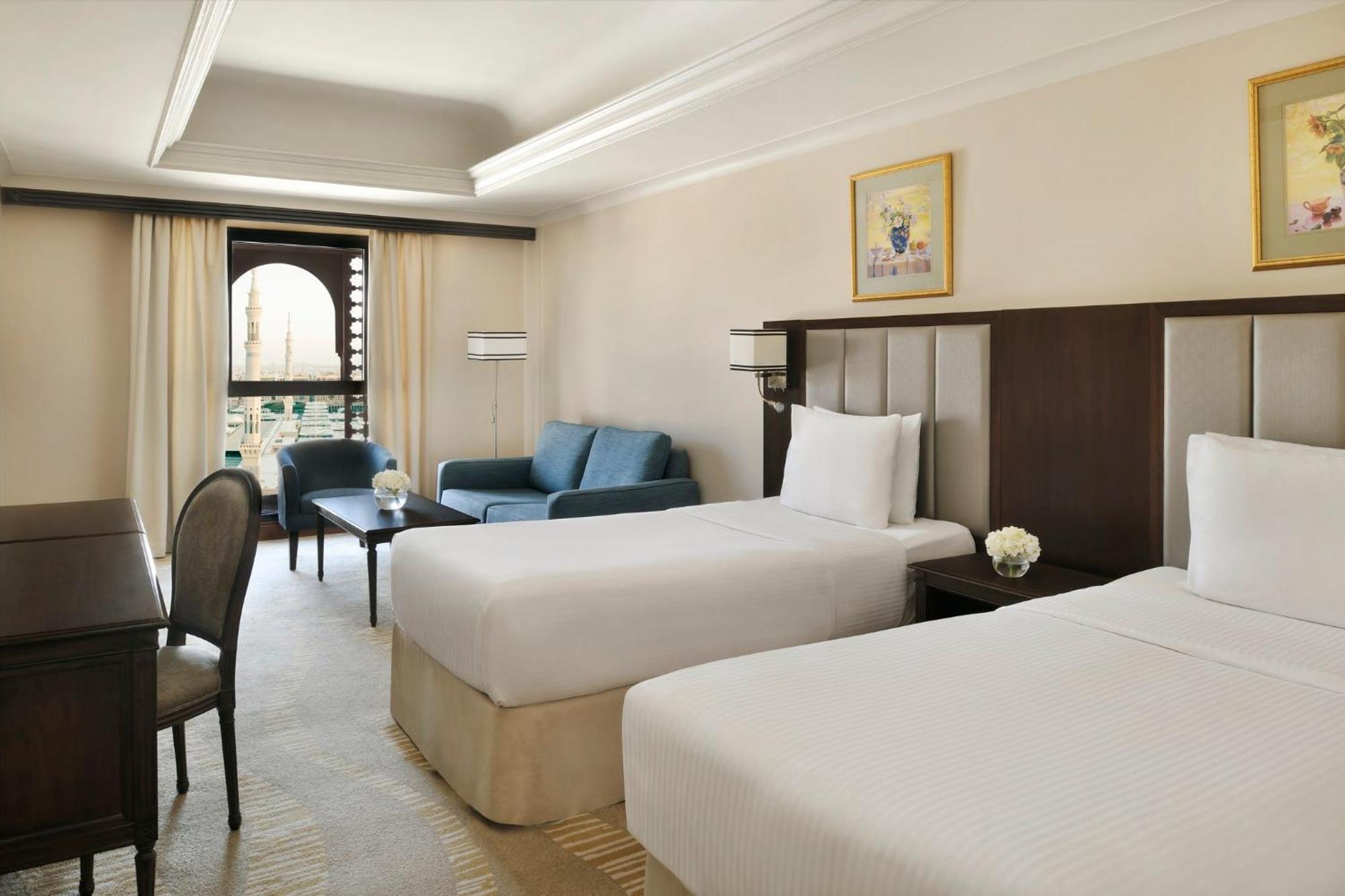 Hotel Intercontinental Dar Al Iman Madinah By Ihg Medyna Zewnętrze zdjęcie The photo shows a well-appointed hotel room featuring two twin beds with white bedding. There is a small desk with a chair, a comfortable blue sofa, and a coffee table in the sitting area. The room has a warm, neutral color scheme, accented by two pa