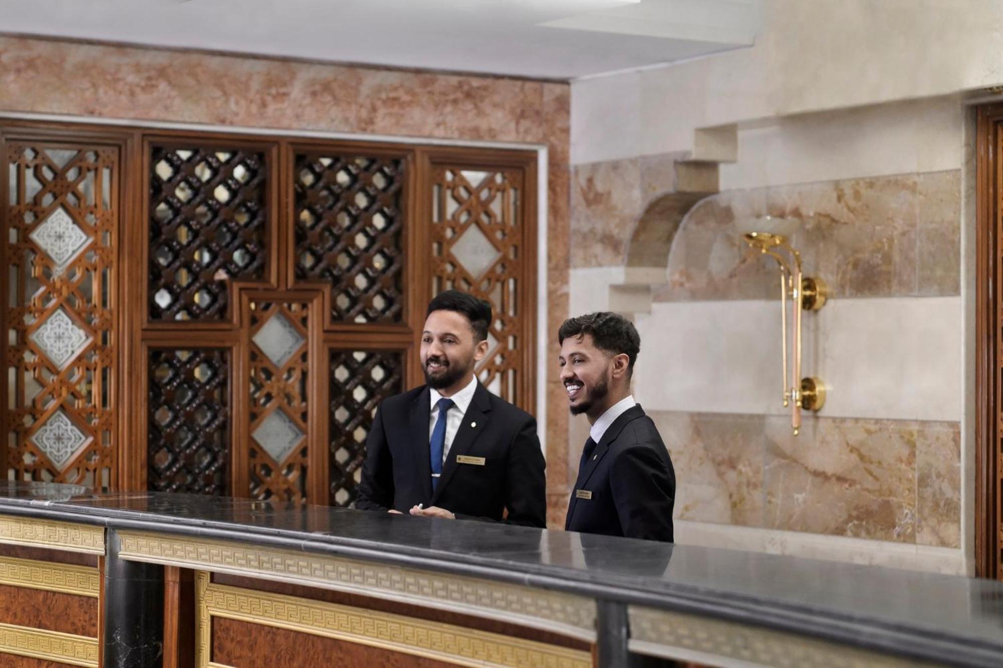 Hotel Intercontinental Dar Al Iman Madinah By Ihg Medyna Zewnętrze zdjęcie The photo shows two men standing behind a reception desk in an elegant setting, likely a hotel or similar establishment. They are both dressed in formal attire, wearing suits and ties, and appear to be smiling or engaging in conversation. The backgro