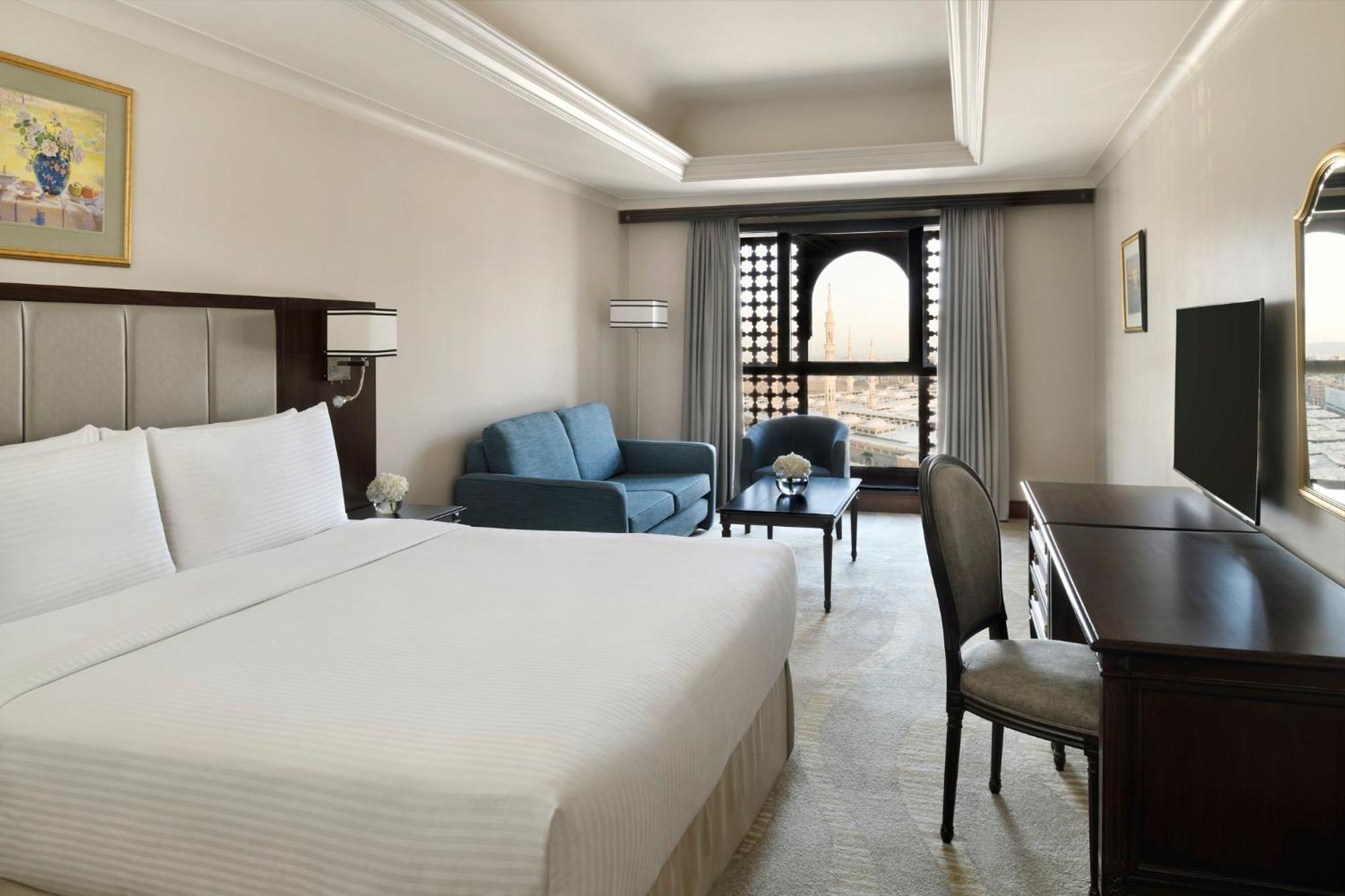 Hotel Intercontinental Dar Al Iman Madinah By Ihg Medyna Zewnętrze zdjęcie The photo shows a hotel room designed with a modern and comfortable aesthetic. It features a large bed with white linens, a seating area with a blue sofa, and a small table. There is a wooden desk with a chair and a television opposite the bed. Natur