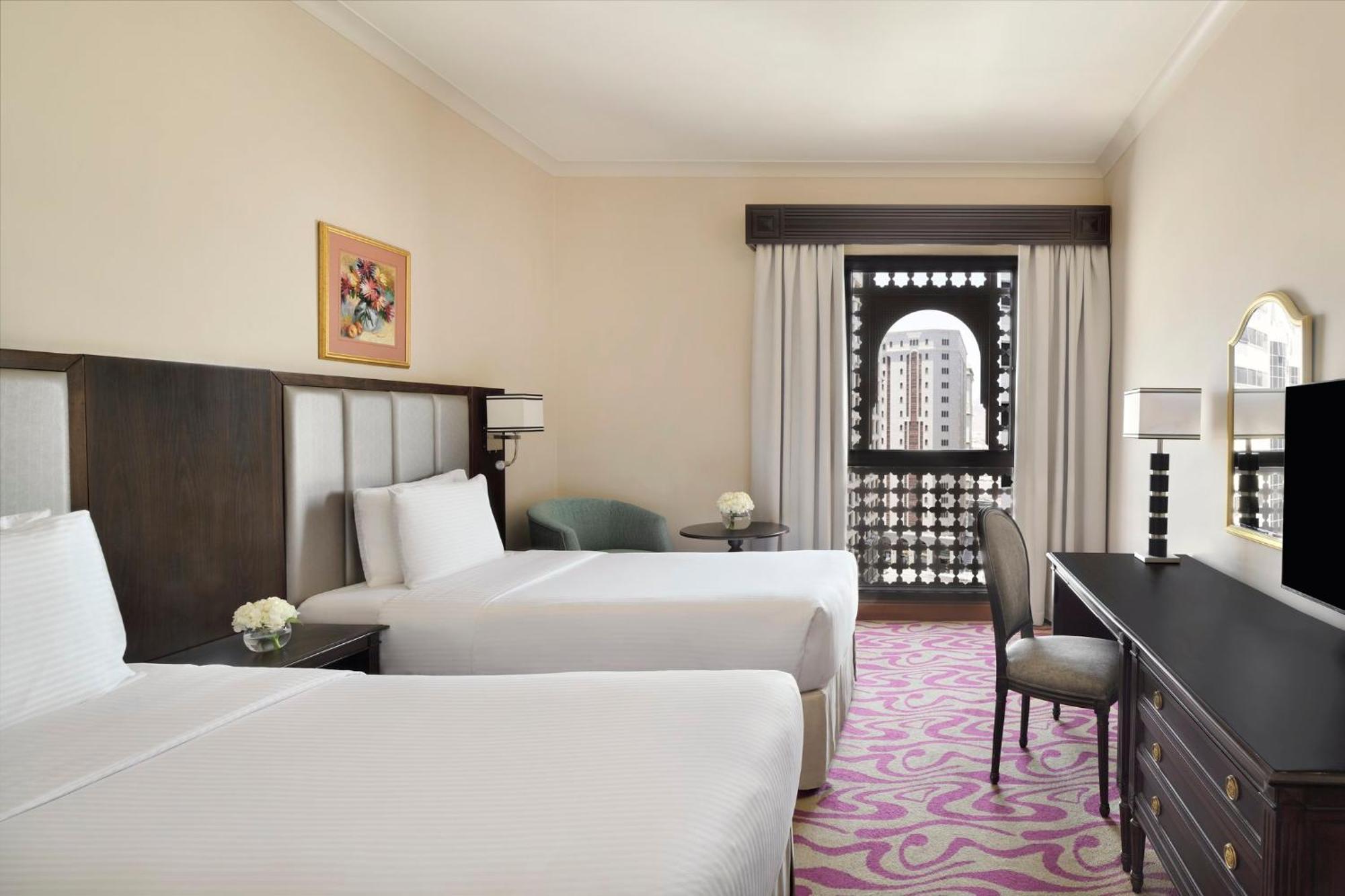 Hotel Intercontinental Dar Al Iman Madinah By Ihg Medyna Zewnętrze zdjęcie The photo shows a hotel room featuring two twin beds with white bedding. There is a decorative piece of art on the wall above one of the beds. A small round table with a flower arrangement is placed next to the beds. A desk and chair are situated aga