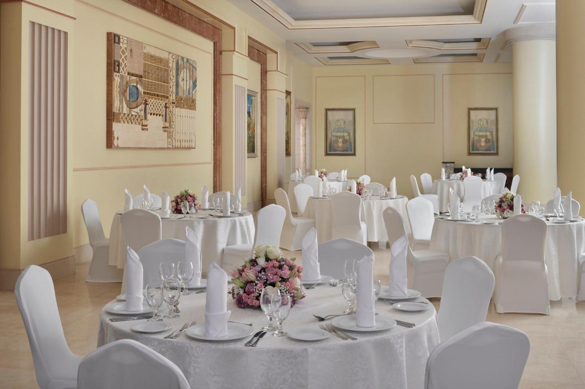 Hotel Intercontinental Dar Al Iman Madinah By Ihg Medyna Zewnętrze zdjęcie The photo shows an elegantly arranged dining or function room. There are round tables covered with white tablecloths, each set with fine dining ware including glassware, utensils, and neatly folded white napkins. Some tables have floral centerpieces 