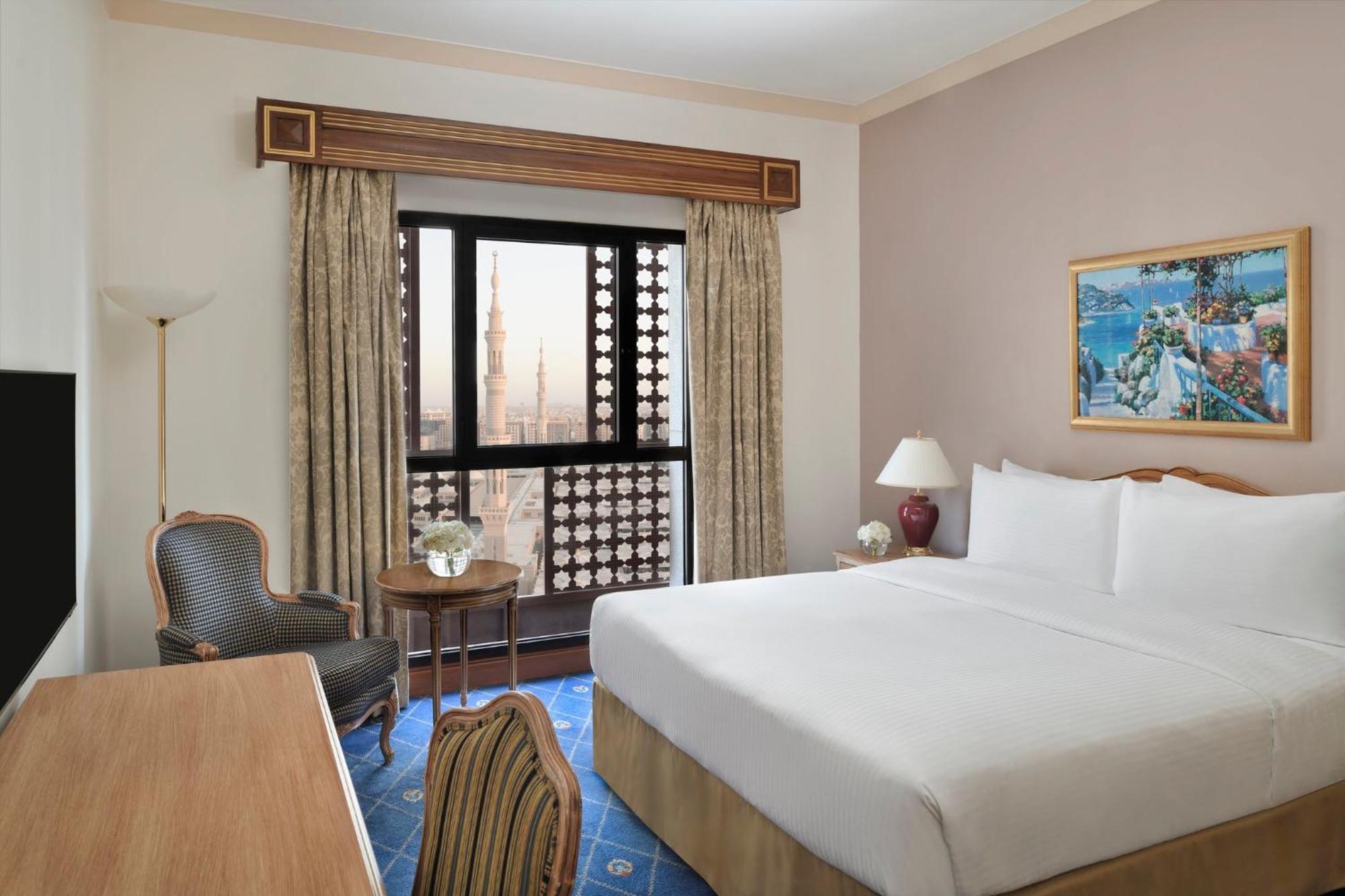 Hotel Intercontinental Dar Al Iman Madinah By Ihg Medyna Zewnętrze zdjęcie The photo shows a hotel room with a modern design. There is a large bed with white linens placed against the wall. Next to the bed, there is a small bedside table with a lamp. A comfortable armchair is situated nearby, accompanied by a wooden desk. T