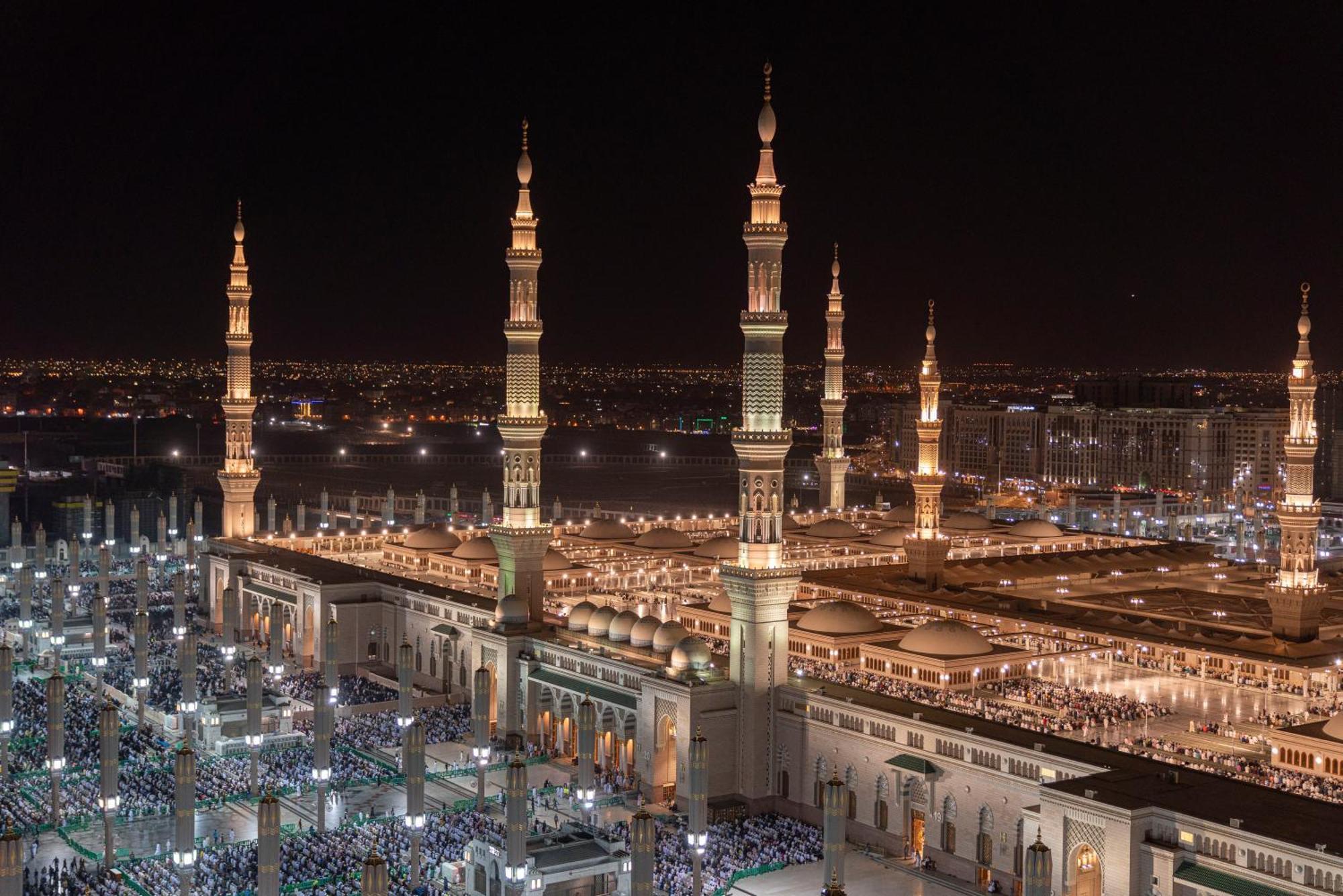 Hotel Intercontinental Dar Al Iman Madinah By Ihg Medyna Zewnętrze zdjęcie The photo depicts a stunning nighttime view of a large mosque complex, prominently featuring tall minarets that are illuminated against the dark sky. The surrounding area is bustling with people, likely worshippers, gathered in the courtyard of the m