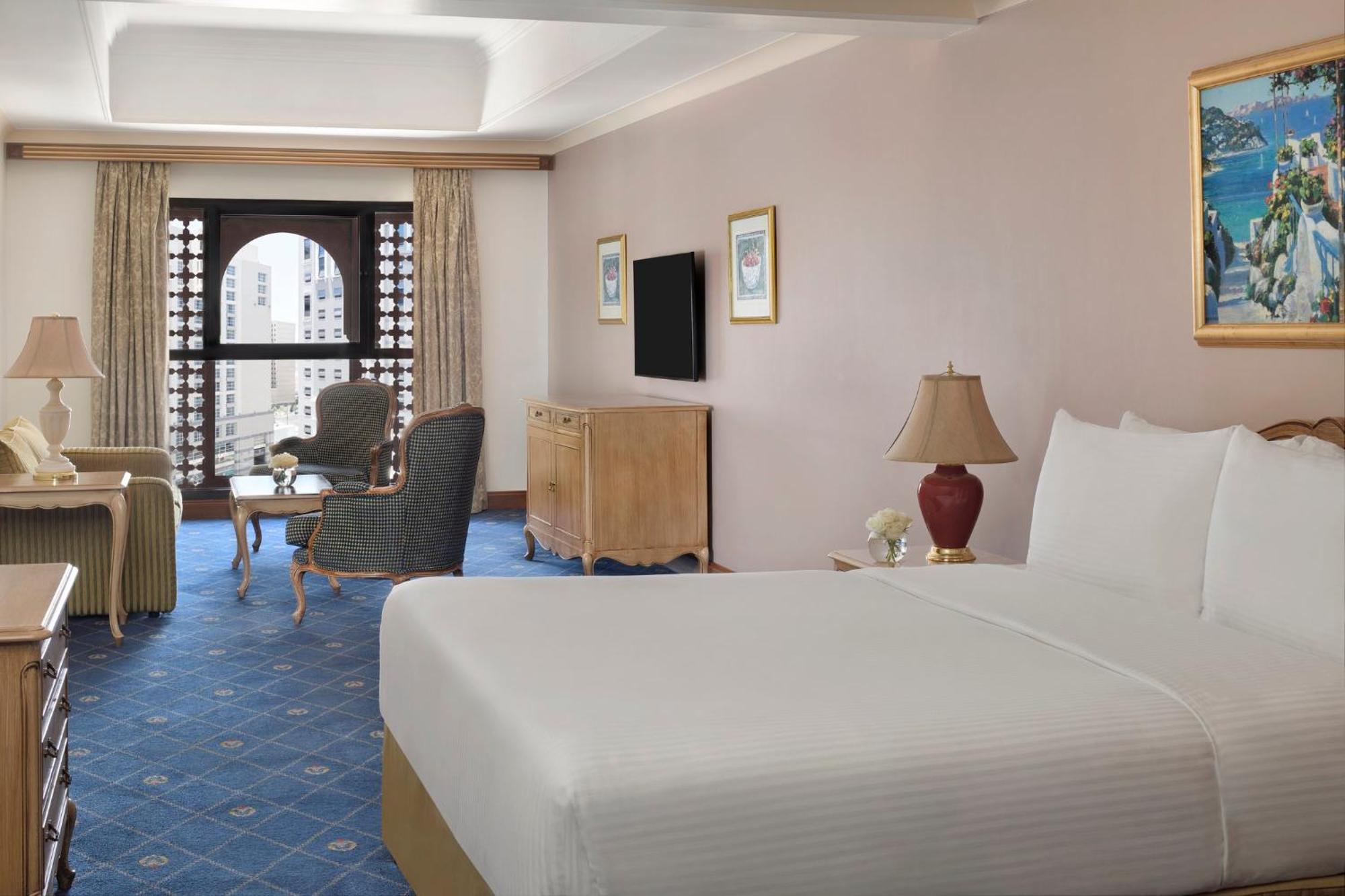 Hotel Intercontinental Dar Al Iman Madinah By Ihg Medyna Zewnętrze zdjęcie The photo shows a well-furnished hotel room. It features a large bed with a white duvet, two bedside lamps, and decorative paintings on the walls. There is a comfortable seating area with two chairs and a small table. A wooden dresser is positioned a