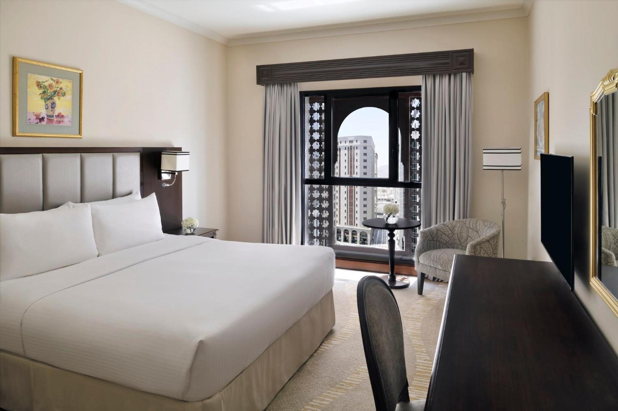 Hotel Intercontinental Dar Al Iman Madinah By Ihg Medyna Zewnętrze zdjęcie The photo shows a hotel room with a modern and minimalistic design. It features a large bed with white linens, a side table, and a comfortable chair near a window. The window has a view of a cityscape, and elegant curtains frame the window. There is 