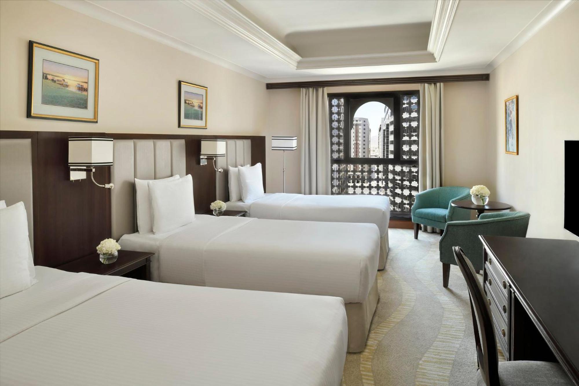 Hotel Intercontinental Dar Al Iman Madinah By Ihg Medyna Zewnętrze zdjęcie The photo shows a hotel room with three single beds, dressed in white linens. There are two paintings on the walls, depicting serene landscapes. A large window allows natural light to enter, and it is adorned with elegant curtains. A smaller seating 