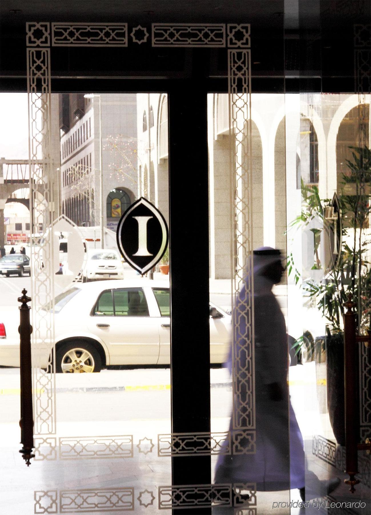Hotel Intercontinental Dar Al Iman Madinah By Ihg Medyna Wnętrze zdjęcie The photo shows a view through a glass door or window, featuring an elegant design with a prominent "I" symbol displayed. Through the glass, there are some parked vehicles visible, including a white car. In the foreground, a shadowy figure of a perso