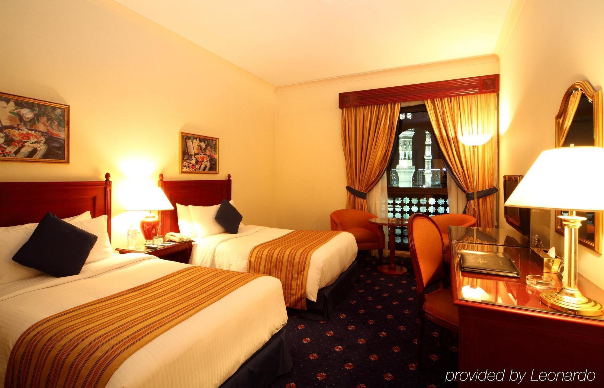 Hotel Intercontinental Dar Al Iman Madinah By Ihg Medyna Pokój zdjęcie The photo shows a hotel room featuring two twin beds with white linens and brown striped bedspreads. There are decorative pillows on the beds, and the room has warm-colored walls. To the side, there's a small sitting area with a round table and a cha