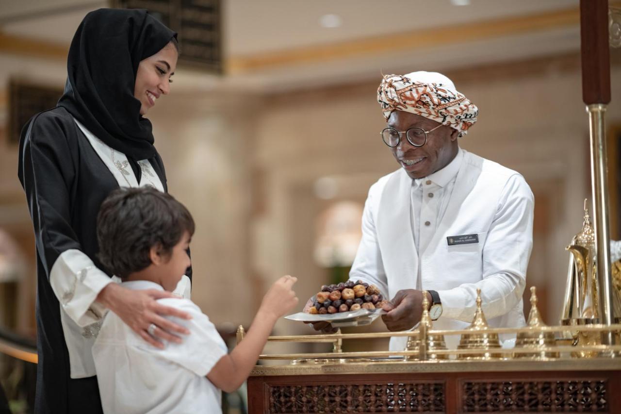 Hotel Intercontinental Dar Al Iman Madinah By Ihg Medyna Zewnętrze zdjęcie The photo depicts a warm interaction between three people. A woman, dressed in a traditional black attire and a headscarf, stands beside a young boy who is reaching out towards a plate of dates held by a smiling man in a white traditional outfit with